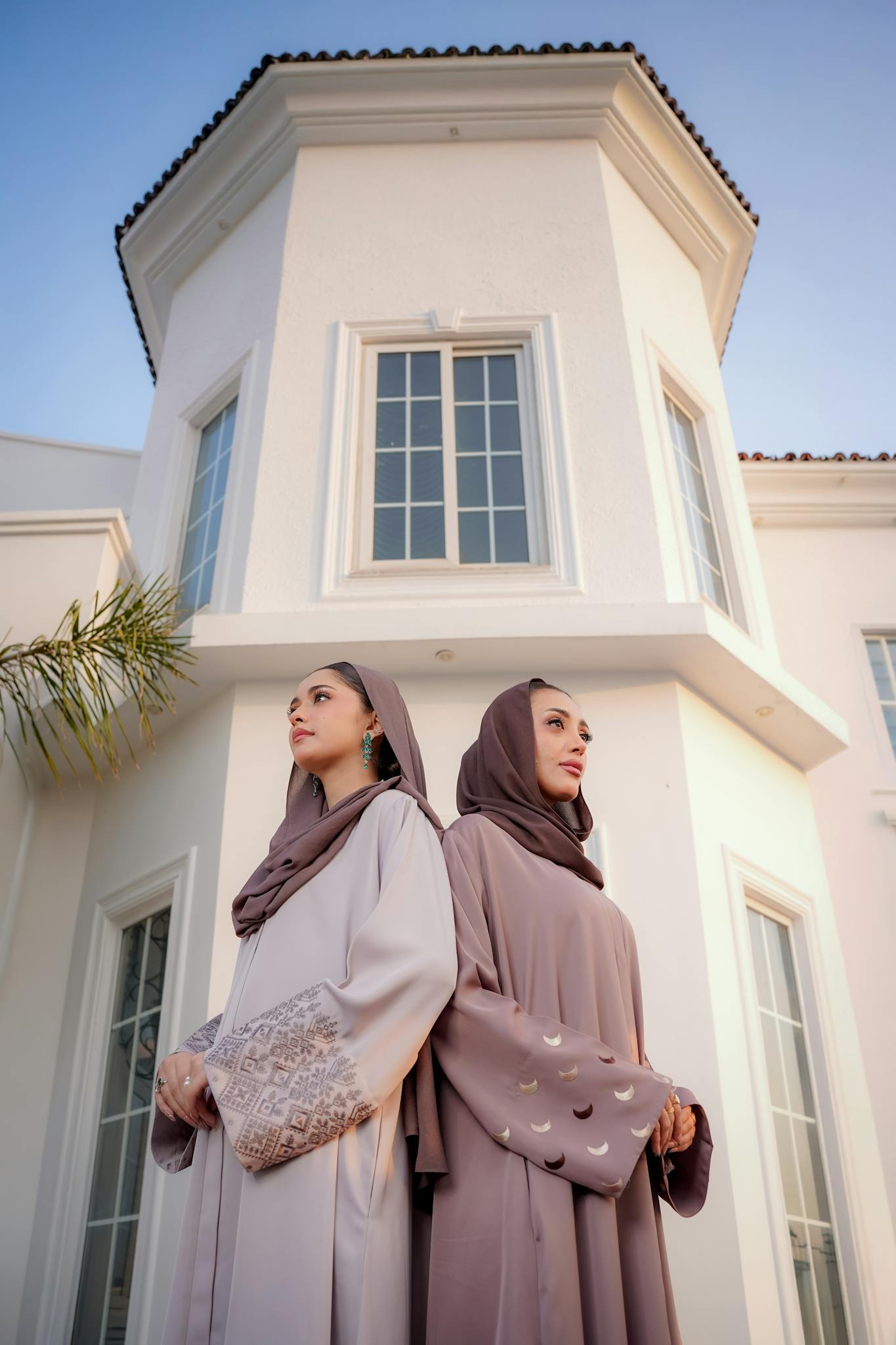 Two women in stylish hijabs and abayas pose elegantly outdoors near a modern building.