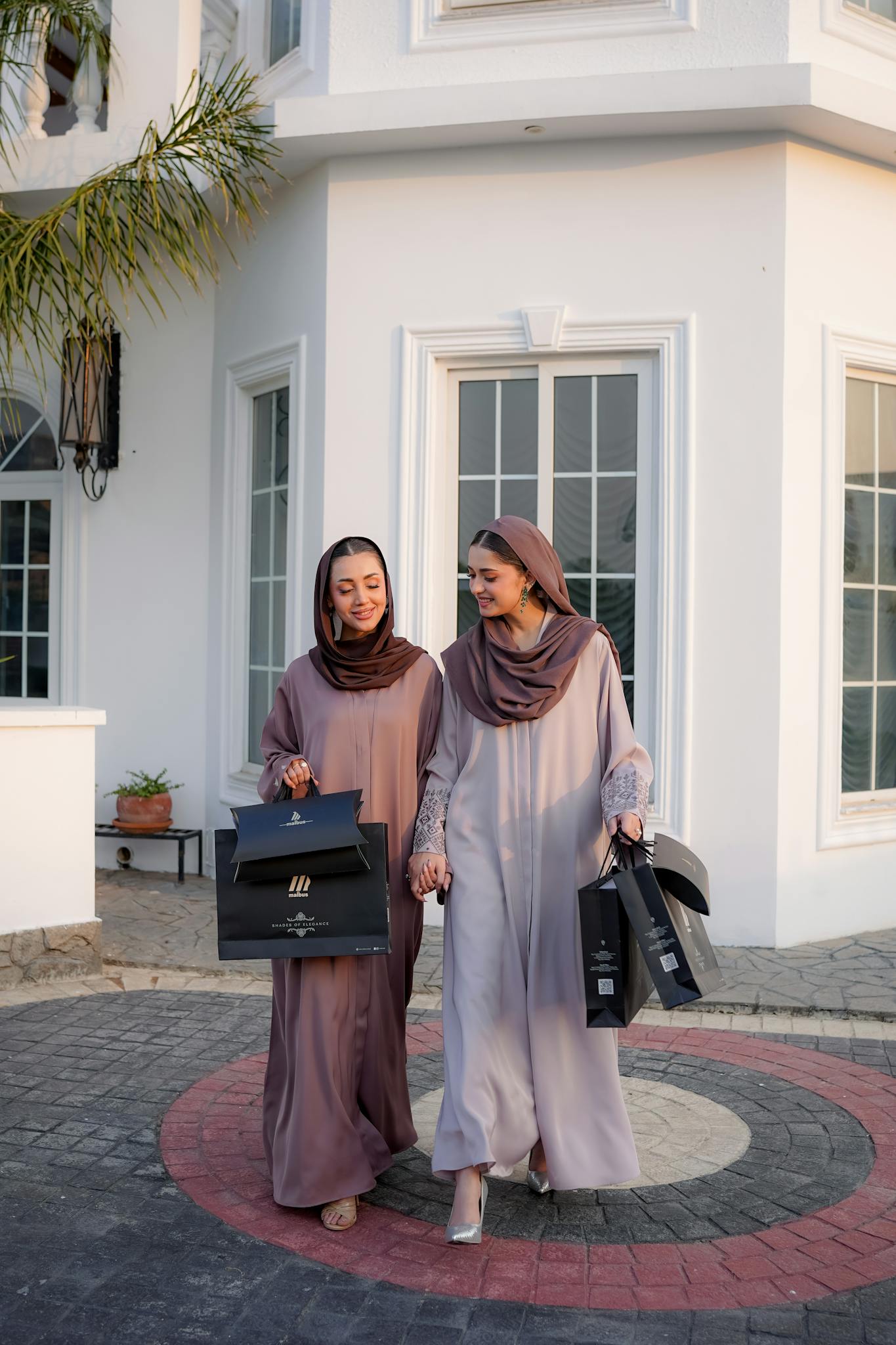 Two women wearing hijabs and modest fashion outfits carrying shopping bags outdoors.