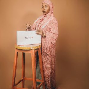 Young woman in a pink hijab holding a 'Thank You' bag indoors against a brown background.
